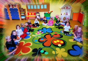 Grupa dzieci siedzi na dywanie. Przed nimi leżą zielone obręcze, w których znajdują się kolorowe koła.