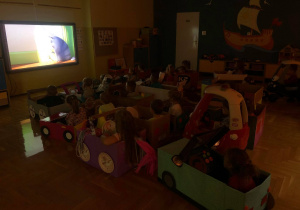 Dzieci w swoich autach, w zacienionej sali oglądają bajkę na tablicy multimedialnej.