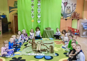 Dzieci na dywanie na tle dekoracji, na środku wykonana makieta drzewa.