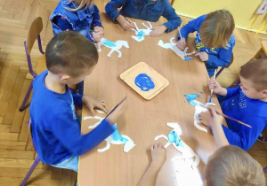 Dzieci przy stoliczkach malują konika niebieską farbą.