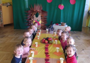 Dzieci siedzą przy jesiennie przystrojonym stoliku, na którym znajduje się taca z owocami