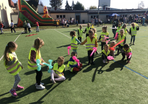 Dzieci z balonami pozwijanymi w różne kształty siedzą na sztucznej strawie boiska przy szkole