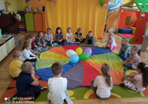 Dzieci siedzą dookoła kolorowej chusty z balonami.