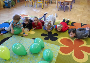 Dzieci leżą na dywanie i dmuchają na zielone balony.