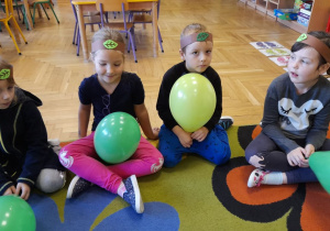 Dziewczynki w opaskach na głowie, siedzą na dywanie i trzymają w rączkach balony.