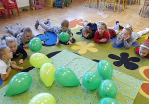 Dzieci leżą na dywanie, dmuchają na swoje baloniki.