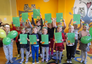 Grupa dzieci w sali przedszkolnej trzyma w rączkach literki tworzące napis "Dzień Drzewa"